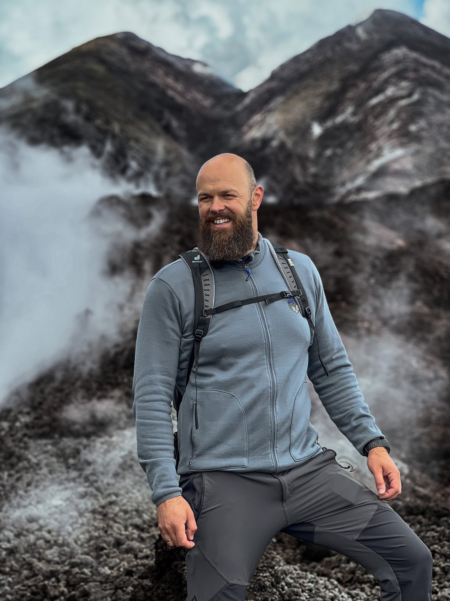 Mann in einer grauen Merino-Jacke mit einem Rucksack steht vor einem schönen Berg