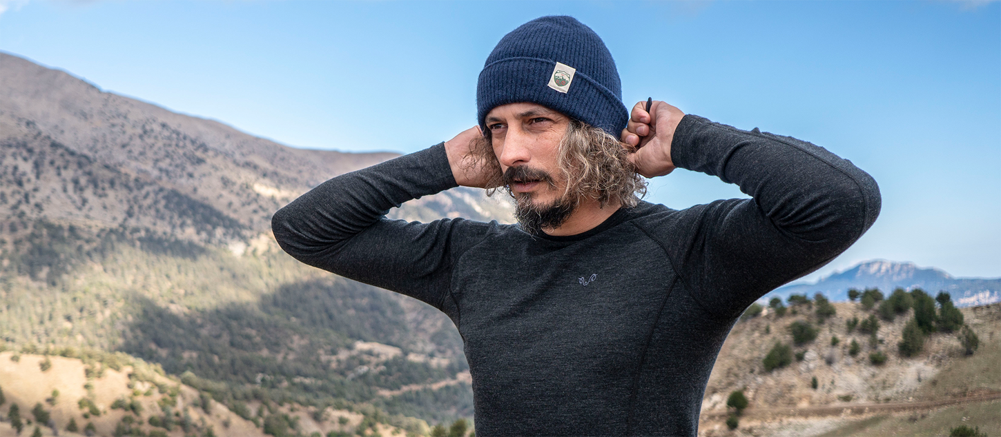 Bärtiger Mann trägt Merino Funktionskleidung und Mütze und steht in einer bergigen Landschaft